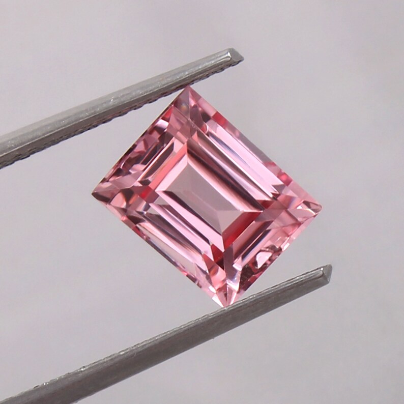 May include: A pink gemstone with a rectangular emerald cut. The gemstone is held by a pair of tweezers.