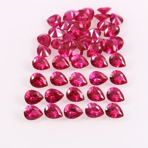 Pode incluir: Uma coleção de pedras preciosas de rubi em forma de pêra, dispostas em filas sobre uma superfície branca. Os rubis são de cor vermelha intensa e têm um alto nível de brilho.
