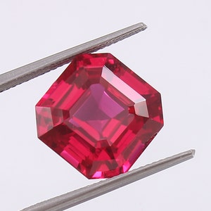 May include: A deep red gemstone with a square emerald cut. The gemstone is held in place by a pair of tweezers.