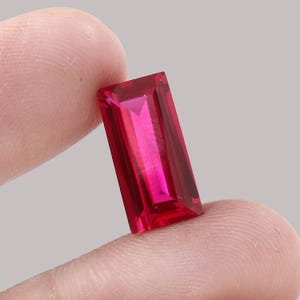 May include: A vibrant, rectangular-cut ruby gemstone. The gem is a deep, saturated red with facets that catch the light. The ruby is held between fingers, showcasing its clarity and brilliance. Ideal for jewelry making.