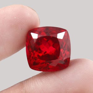 May include: A faceted, cushion-cut, red gemstone. The gem is held between two fingers against a light gray background. The gem's facets reflect light, creating a brilliant display of color and sparkle. The gemstone is a deep, rich red.