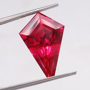 Może przedstawiać: Czerwony kamień szlachetny o głębokim kolorze, o fasetowanym, latawcowatym szlifie. Kamień szlachetny jest trzymany przez pęsetę.