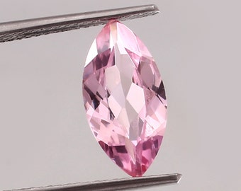 Puede incluir: Una piedra preciosa rosa de talla marquesa sostenida por unas pinzas. La piedra preciosa está facetada y tiene un alto nivel de claridad.