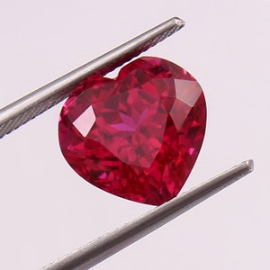 May include: A heart-shaped ruby gemstone with a deep red color. The gemstone is held in a pair of tweezers.