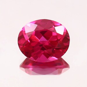 May include: A vibrant, oval-shaped ruby gemstone with a deep red hue and a faceted cut. The gemstone is set against a white background.