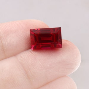 Puede incluir: Una piedra preciosa rectangular de color rojo intenso con un corte facetado. La piedra preciosa se sostiene en una mano.