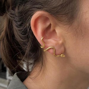 Puede incluir: Un pendiente tipo ear cuff de color dorado con forma de serpiente esbelta y sinuosa. La pieza presenta una superficie texturizada similar a escamas que se curva a lo largo del borde exterior de la oreja, con la cabeza cerca del lóbulo.