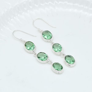 May include: A pair of silver earrings with three oval, light green gemstones each. The gemstones are set in a silver bezel and dangle from a silver hook. The earrings are displayed on a white surface.