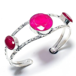 May include: A silver cuff bracelet featuring three vibrant, faceted, oval and round, hot pink gemstones. The bracelet has an open design with ornate, etched detailing along the band. The gemstones are set in a silver bezel.