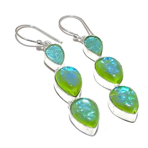 Könnte beinhalten: Silberne Hängeohrringe mit drei tropfenförmigen, irisierenden grün-blauen Steinen. Die Ohrringe haben einen Hakenverschluss und sind in Silber gefasst. Die Steine haben eine lebendige, auffällige Farbe.