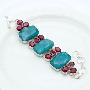 May include: A silver bracelet featuring three large, rectangular, faceted teal stones alternating with clusters of small, round, deep red rubies. The piece includes a silver toggle clasp and a series of circular links for adjustable sizing.