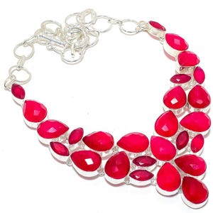 Puede incluir: Un collar plateado con una serie de gemas rojas brillantes, facetadas, en forma de lágrima y ovalada. Las gemas están engastadas en biseles individuales de plata y conectadas por una cadena de plata.