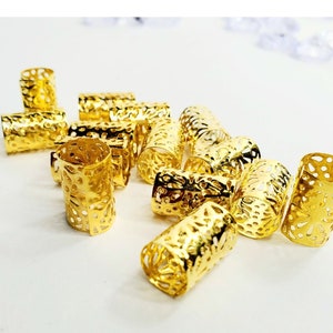 May include: A group of gold-colored metal hair beads with a decorative, openwork design. The beads are scattered on a white surface.