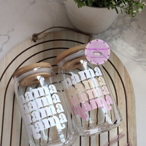 May include: Two clear glass tumblers with bamboo lids and pink and white "mama" text. The tumblers are on a wooden surface with two pink straws.