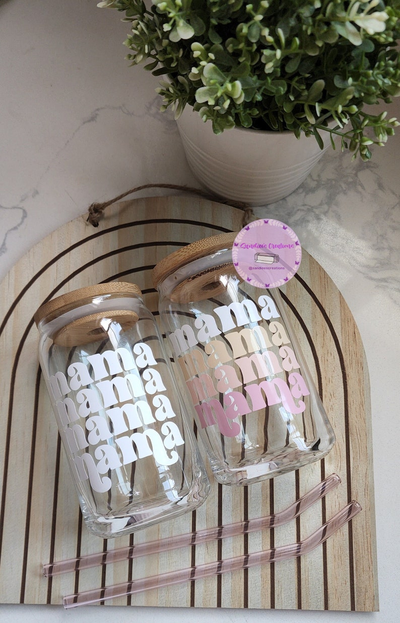 May include: Two clear glass tumblers with bamboo lids and pink and white "mama" text. The tumblers are on a wooden surface with two pink straws.