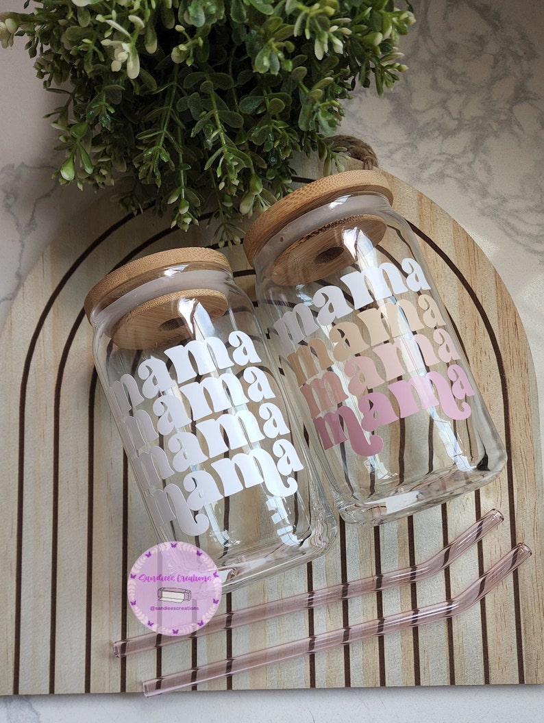 May include: Two clear glass jars with bamboo lids and pink and white "mama" text printed on them. The jars are sitting on a wooden surface with two pink glass straws.