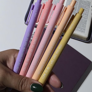 May include: A hand holds five pastel-colored pens with inspirational quotes and Bible verses printed on them. The pens are in shades of lavender, pink, peach, and yellow. A book and a purple notebook are in the background.