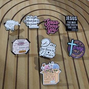 May include: A collection of colorful enamel pins with religious and inspirational themes. Designs include a sheep, floral arrangements, and text such as "Jesus Loves You" and "Consider How the Wild Flowers Grow."