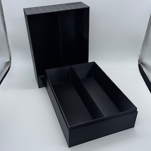 May include: A black box with a lid that opens to reveal two compartments. The box is made of a textured material and has a smooth finish.
