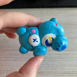 May include: A blue clay figurine of a Care Bear with a white belly and a crescent moon on its chest. The bear has a pink nose and a white X on its eye.