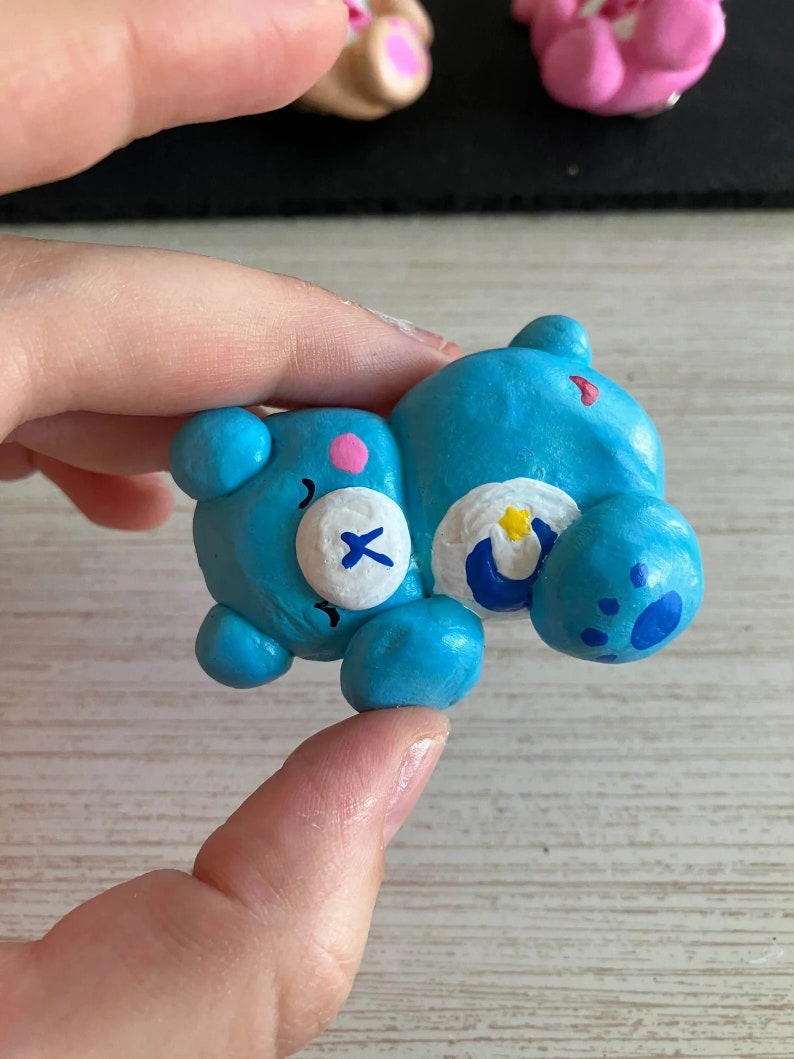 May include: A blue clay figurine of a Care Bear with a white belly and a crescent moon on its chest. The bear has a pink nose and a white X on its eye.