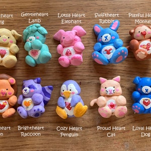 May include: Ten handmade clay figurines of various animals, each with a heart and a name. The animals include a pig, lamb, elephant, rabbit, monkey, lion, raccoon, penguin, cat, and dog. Each figurine is painted in bright colors and has a unique design.