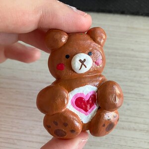 May include: A small brown clay bear figurine with a pink heart on its chest. The bear has a white nose and black eyes.