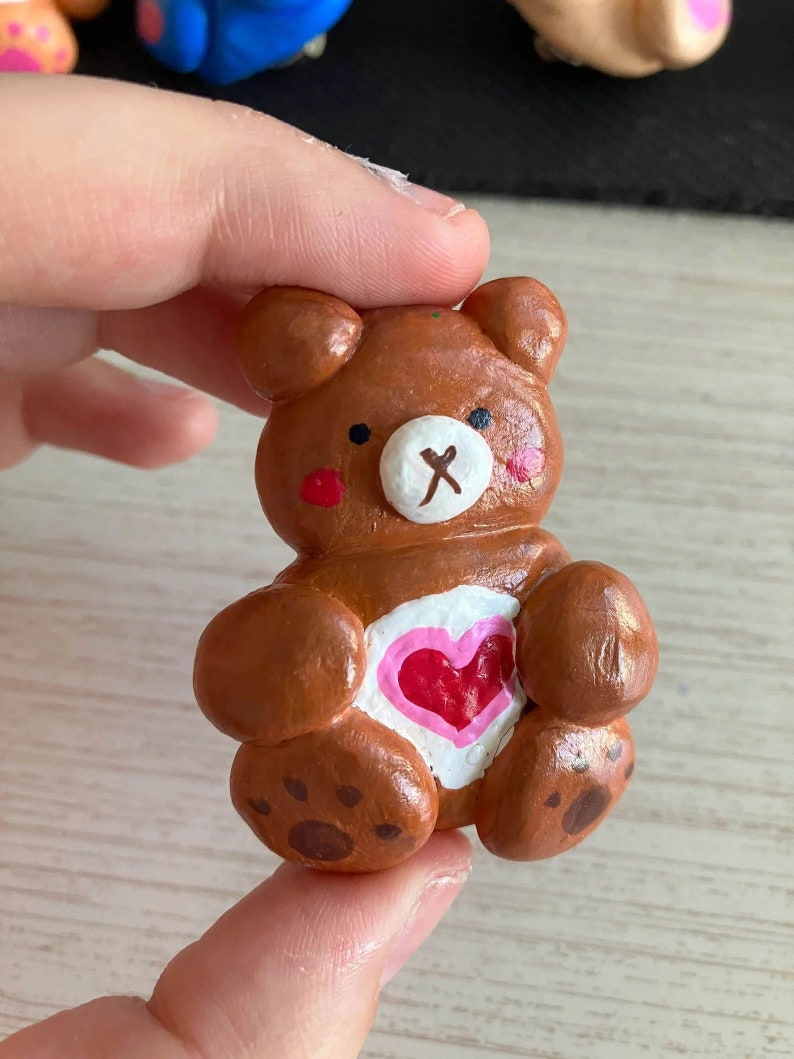 May include: A small brown clay bear figurine with a pink heart on its chest. The bear has a white nose and black eyes.