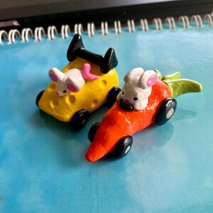 May include: Two whimsical toy cars: one shaped like a yellow cheese block with a white mouse, the other a carrot with a white rabbit. Both have black wheels and a black spoiler. The mouse has pink ears and a pink tail. The rabbit has pink ears.