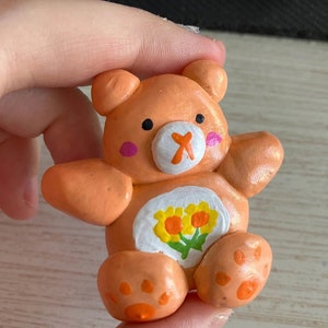May include: A small orange clay bear figurine with a white belly and a flower design. The bear has a painted orange nose, black eyes, and pink cheeks.
