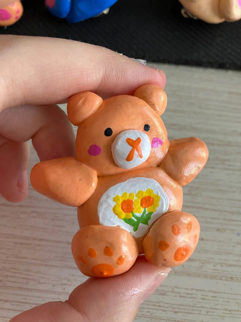 May include: A small orange clay bear figurine with a white belly and a flower design. The bear has a painted orange nose, black eyes, and pink cheeks.