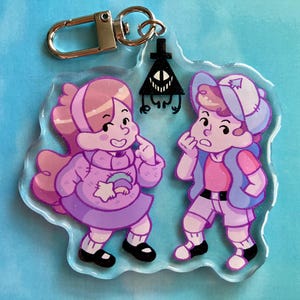 May include: A clear acrylic keychain featuring two cartoon characters in pink and purple outfits. The keychain has a silver clasp and a black triangle with a face. The characters are in a playful pose.