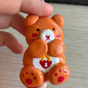 May include: Orange clay bear figurine with a white heart and a lock with the number 1 on it. The bear has red cheeks and a red nose.