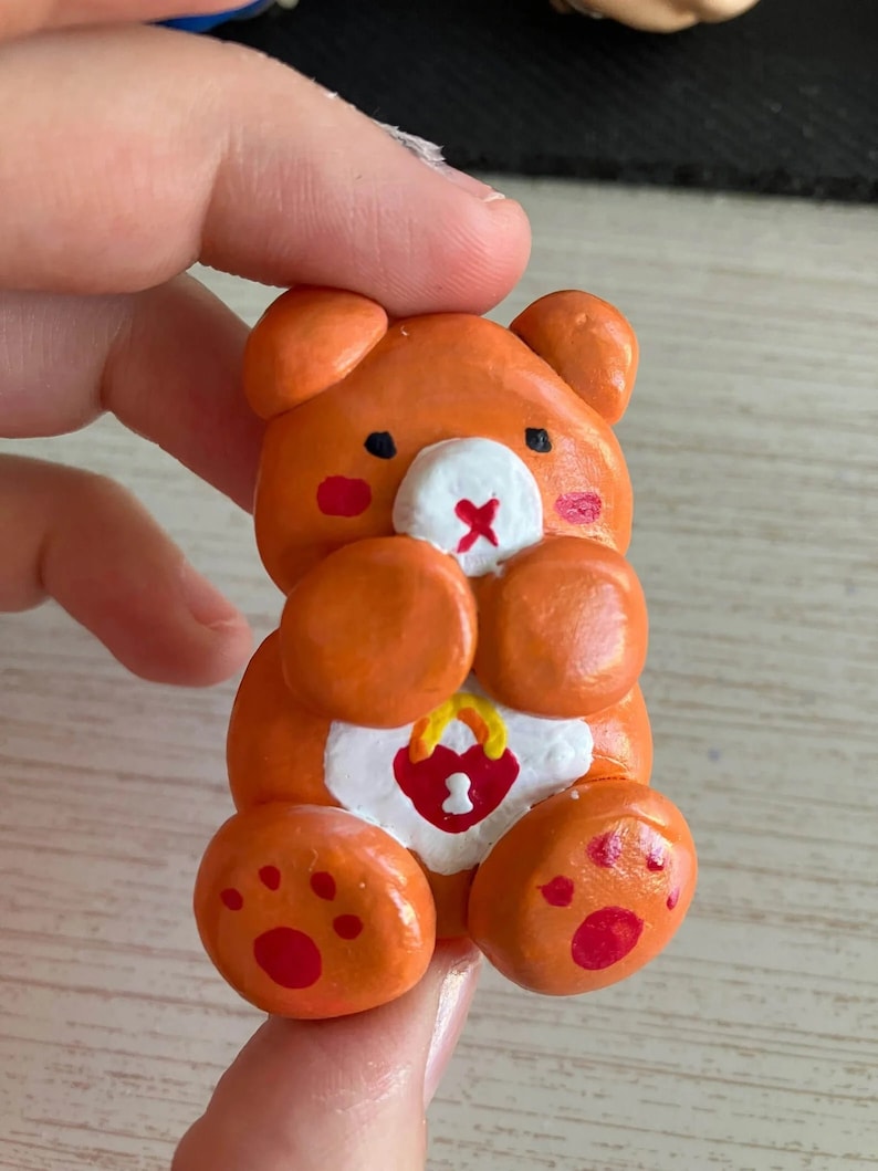 May include: Orange clay bear figurine with a white heart and a lock with the number 1 on it. The bear has red cheeks and a red nose.