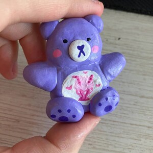 May include: A purple clay figurine of a Care Bear with a pink heart and white wings on its chest. The bear has a white face with black eyes and a white nose with a pink X.