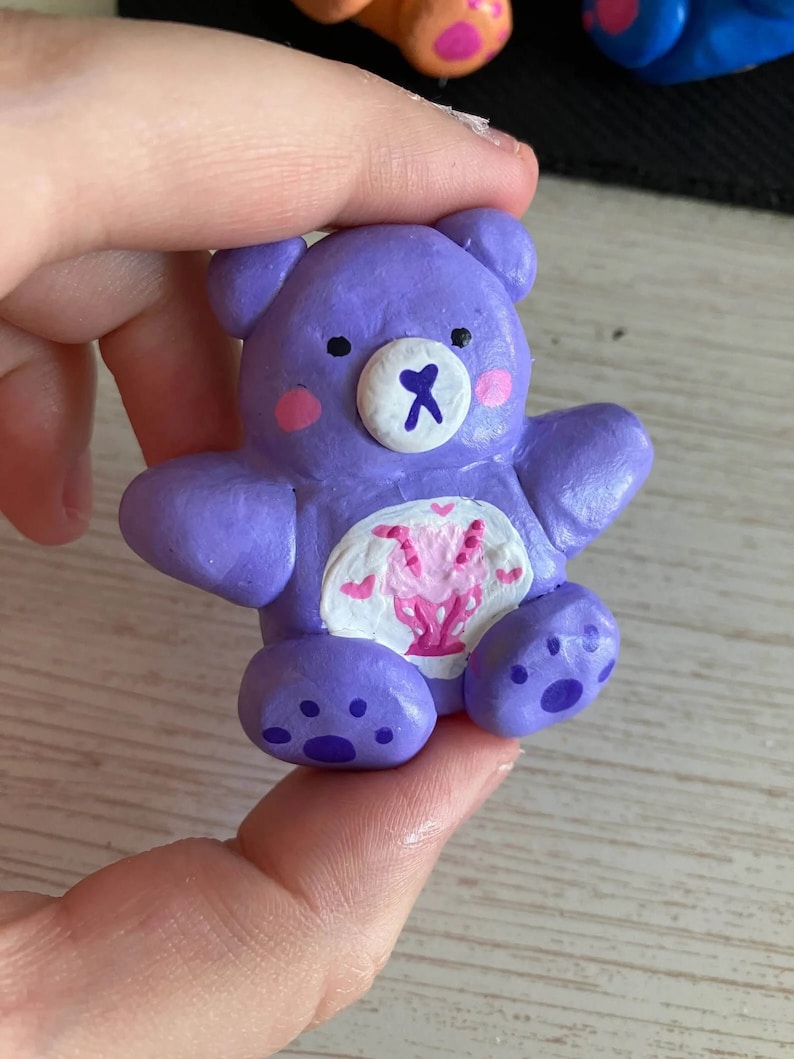 May include: A purple clay figurine of a Care Bear with a pink heart and white wings on its chest. The bear has a white face with black eyes and a white nose with a pink X.