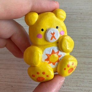 May include: A yellow clay figurine of a bear with a white sun and orange rays on its chest. The bear has a white nose with an orange X on it and pink cheeks.