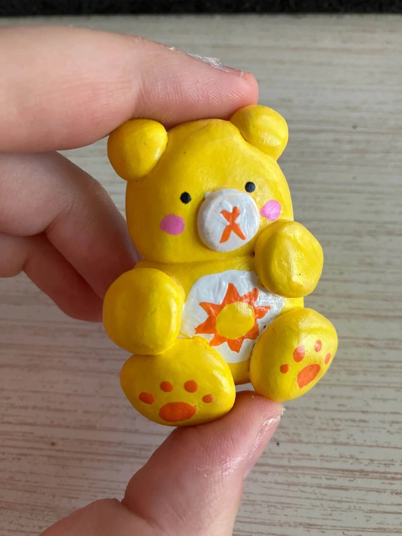 May include: A yellow clay figurine of a bear with a white sun and orange rays on its chest. The bear has a white nose with an orange X on it and pink cheeks.