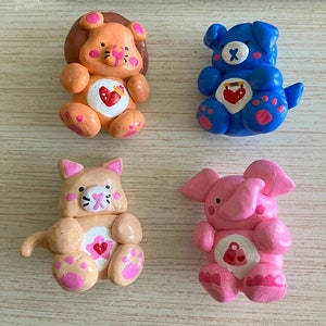 May include: Four handmade clay figurines of Care Bears. The bears are a lion, a dog, a cat, and an elephant. Each bear is wearing a different color and has a different symbol on its chest. The lion is orange, the dog is blue, the cat is peach, and the elephant is pink.