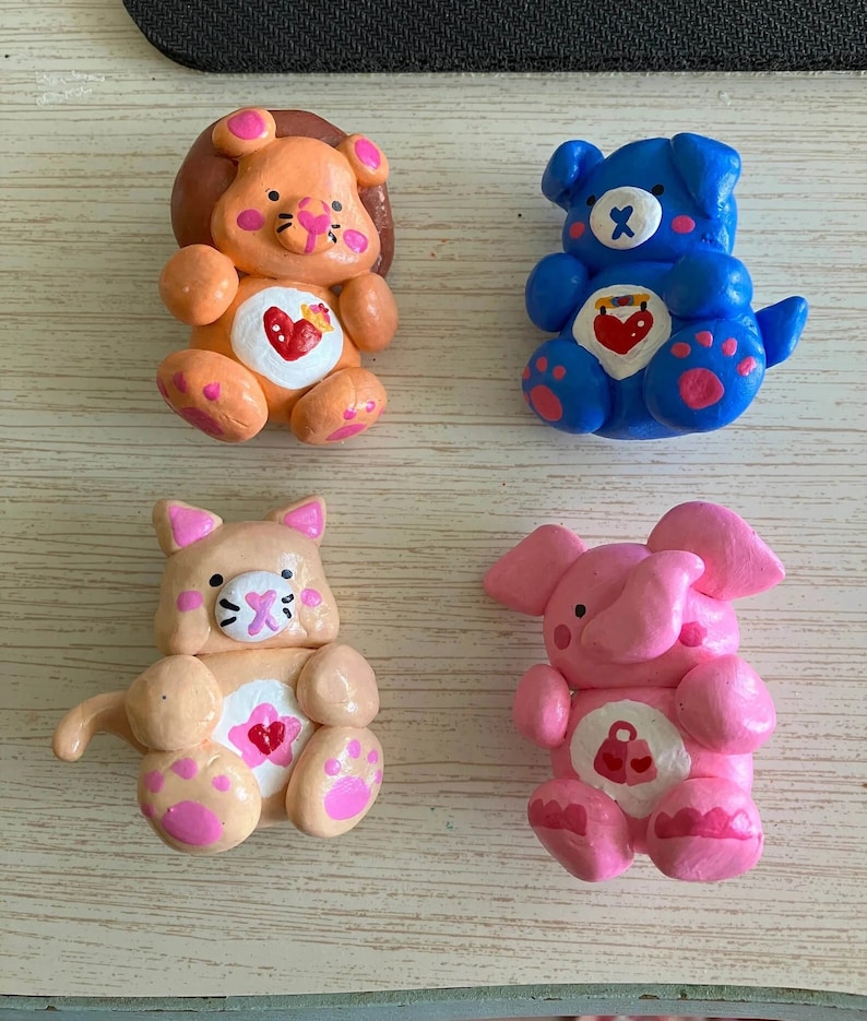 May include: Four handmade clay figurines of Care Bears. The bears are a lion, a dog, a cat, and an elephant. Each bear is wearing a different color and has a different symbol on its chest. The lion is orange, the dog is blue, the cat is peach, and the elephant is pink.