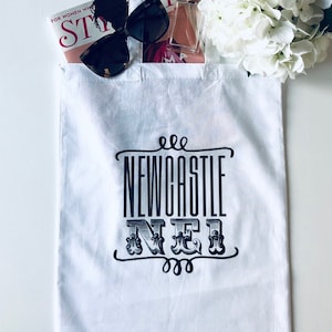May include: White canvas tote bag with black handles. The bag features a black and white graphic design that says "NEWCASTLE NER" with decorative swirls. The bag is filled with a magazine, sunglasses, and a bottle of perfume.