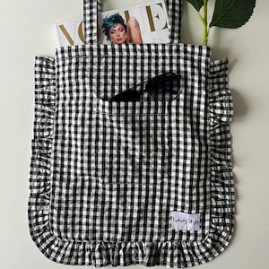 Pode incluir: Uma sacola tote xadrez em preto e branco com uma borda com babados e um bolso. A sacola contém uma revista, óculos de sol e está ao lado de uma flor branca. A sacola tem o texto "Luxury Style" em uma etiqueta.