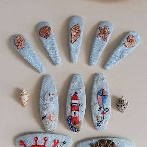 Può includere: Un set di 10 mollette per capelli in tessuto blu con vari disegni ricamati, tra cui conchiglie, una stella marina, un faro, un gabbiano, un aquilone, un granchio, una tartaruga, un arcobaleno e le parole "Summer Love".