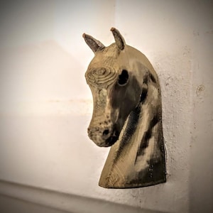 May include: A bronze-colored horse head sculpture mounted on a white wall. The sculpture features detailed textures and a realistic depiction of a horse's head and neck, with visible ears and facial features.