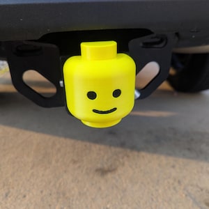 May include: A vibrant yellow Lego-style hitch cover featuring a smiling face. It's attached to a black trailer hitch on a vehicle. This hitch cover provides a fun, playful aesthetic to the vehicle's exterior.