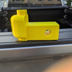 May include: A bright yellow trailer hitch receiver, designed to resemble a toy brick. The hitch features a rectangular body with a circular hole and a brick-like head. It is resting on a truck bed.