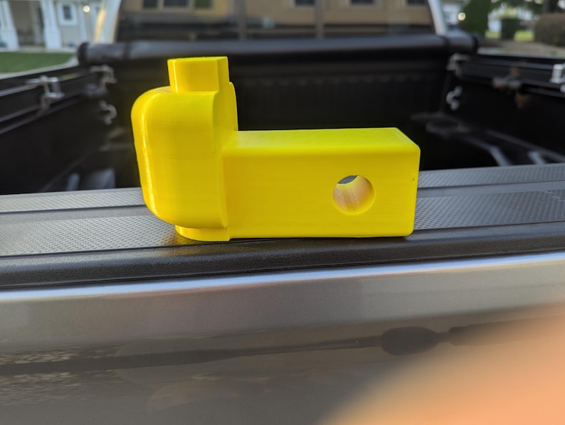 May include: A bright yellow trailer hitch receiver, designed to resemble a toy brick. The hitch features a rectangular body with a circular hole and a brick-like head. It is resting on a truck bed.