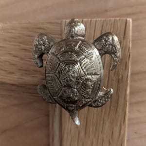 Może przedstawiać: Biały ceramiczny kubek z czarnym uchwytem i napisem "But First, Coffee" pogrubioną, czarną czcionką. Kubek jest ustawiony na rozmytym tle, sugerującym przytulne otoczenie.