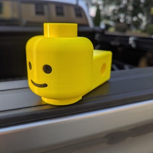 May include: A bright yellow, 3D-printed Lego head with black eyes and a smile. The head is attached to a rectangular base with a hole. The toy is sitting on a black surface.