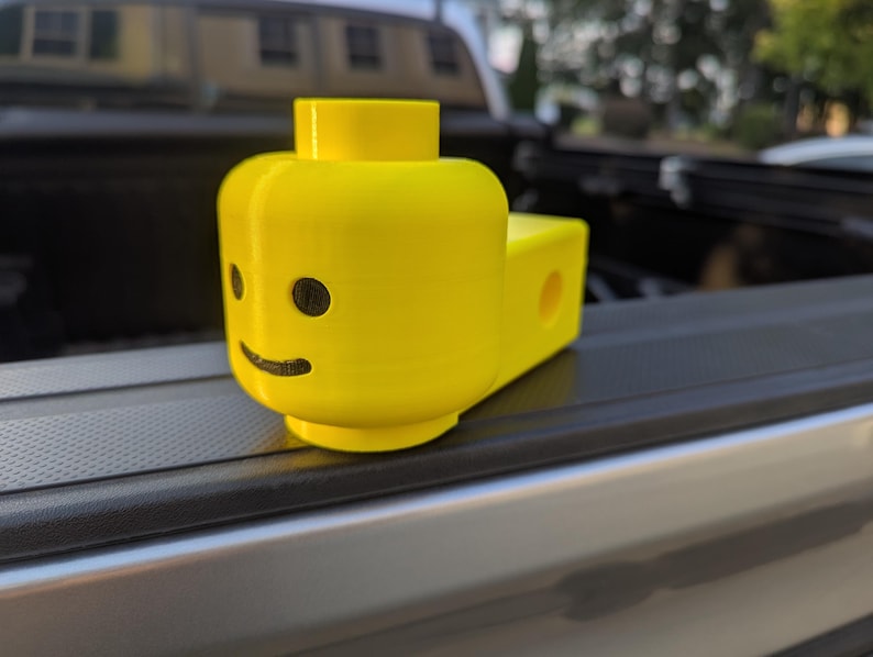 May include: A bright yellow, 3D-printed Lego head with black eyes and a smile. The head is attached to a rectangular base with a hole. The toy is sitting on a black surface.